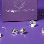 A pair of Aperture Earrings sit on a purple table. In the background is a purple Montgomery-Ramsay product box, and there are some clear gems scattered about. 