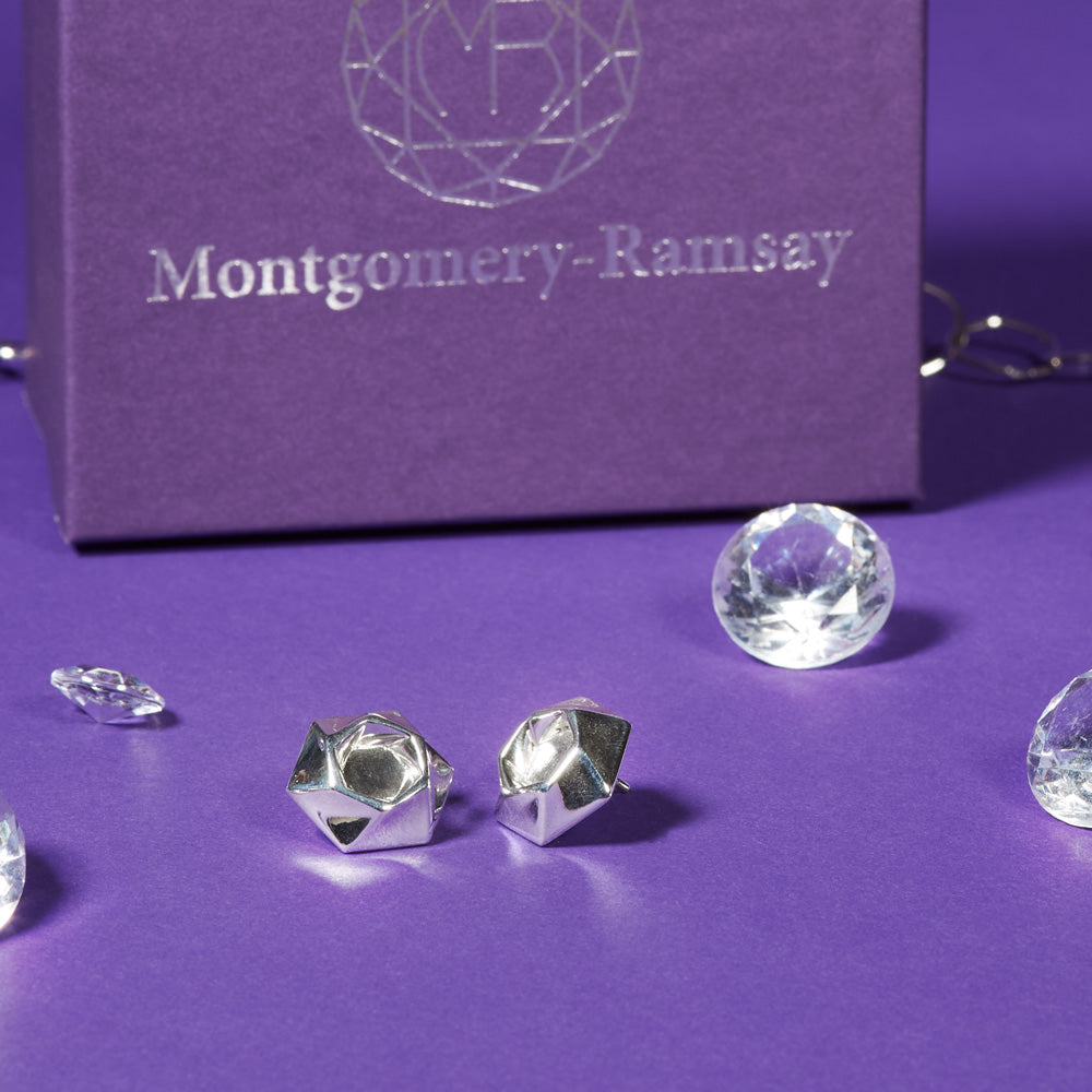 A pair of Aperture Earrings sit on a purple table. In the background is a purple Montgomery-Ramsay product box, and there are some clear gems scattered about. 