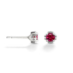A pair of small silver studs on a white background. The gems at the center of each stud are a deep pink tourmaline. One of the earrings is shown in a side-view, revealing the small heart designs on the sides of each. 