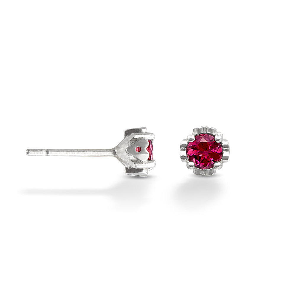 A pair of small silver studs on a white background. The gems at the center of each stud are a deep pink tourmaline. One of the earrings is shown in a side-view, revealing the small heart designs on the sides of each. 