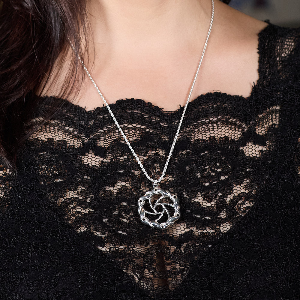 The image shows a close-up of a model wearing a black lace top. The focus is on the Maelstrom Necklace she is wearing. The necklace hangs on a rope chain, complementing the pendant. The background is blurred, emphasizing the necklace and the lace top. The overall aesthetic is elegant and sophisticated.