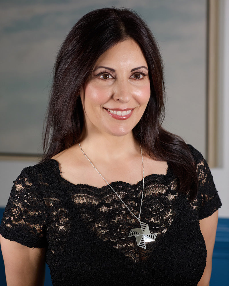 A photo of a brunette model with slightly longer than shoulder length hair, wearing the Windmolen Necklace over a lace black top. The model is smiling and looking over the right shoulder of the photographer. The background is blurred. 