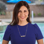 A photo of a smiling, brunette model with shoulder length hair, wearing the Zeta Corridor Necklace. The necklace contrasts nicely on her royal blue shirt, and you can see an our of focus swimming pool in the background. 