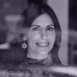 A monochrome image taken through a window of a model wearing the atom earrings. The model is a brunette with shoulder-length hair, and is smiling at the camera.