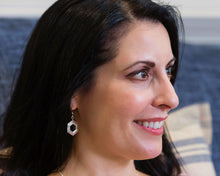 Load image into Gallery viewer, A smiling brunette model wearing our Stepped Hexagon Earrings. The model is looking toward the right of the frame, and we&#39;re looking at the earring hanging from the model&#39;s right ear. It&#39;s indoors, and there is a blue couch behind her. 
