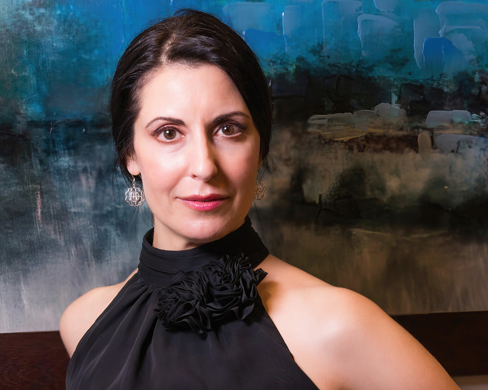 A photo from the shoulders up of a brunette model wearing our Supernova Earrings. The model is staring at the camera. The photo was taken indoors, and there is art in the background. 