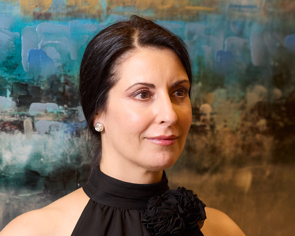 A brunette model wears our Rosette Earrings. The model is looking off to photo right, and is wearing a black top. She has a slight smirk, but is otherwise looking serious. The background is a painting with blues and tans. 