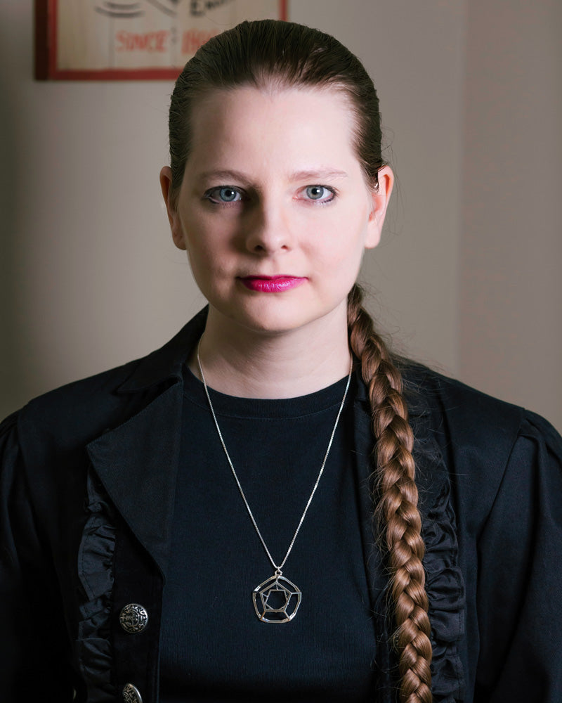 A photo of a blonde model wearing our Dodecahedron Necklace. The model is indoors, wearing black, and has a long blonde braid hanging across the left side of her body.