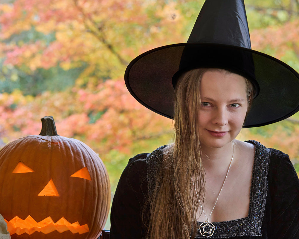 A photo of a model dressed as a witch wearing our Dodecahedron Necklace. The model is sitting next to a jack-o-lantern and the background is out of focus Autumn foliage. 