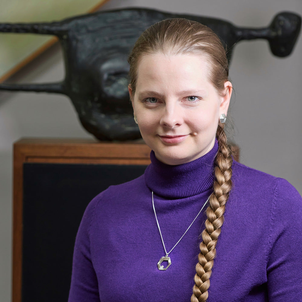 A blonde model in a purple turtleneck wears a Hextech Neclace. She is smiling and looking at the camera. Her long, blonde braid hangs down the front of her turtleneck next to the necklace. In the background is blurred sculpture. 