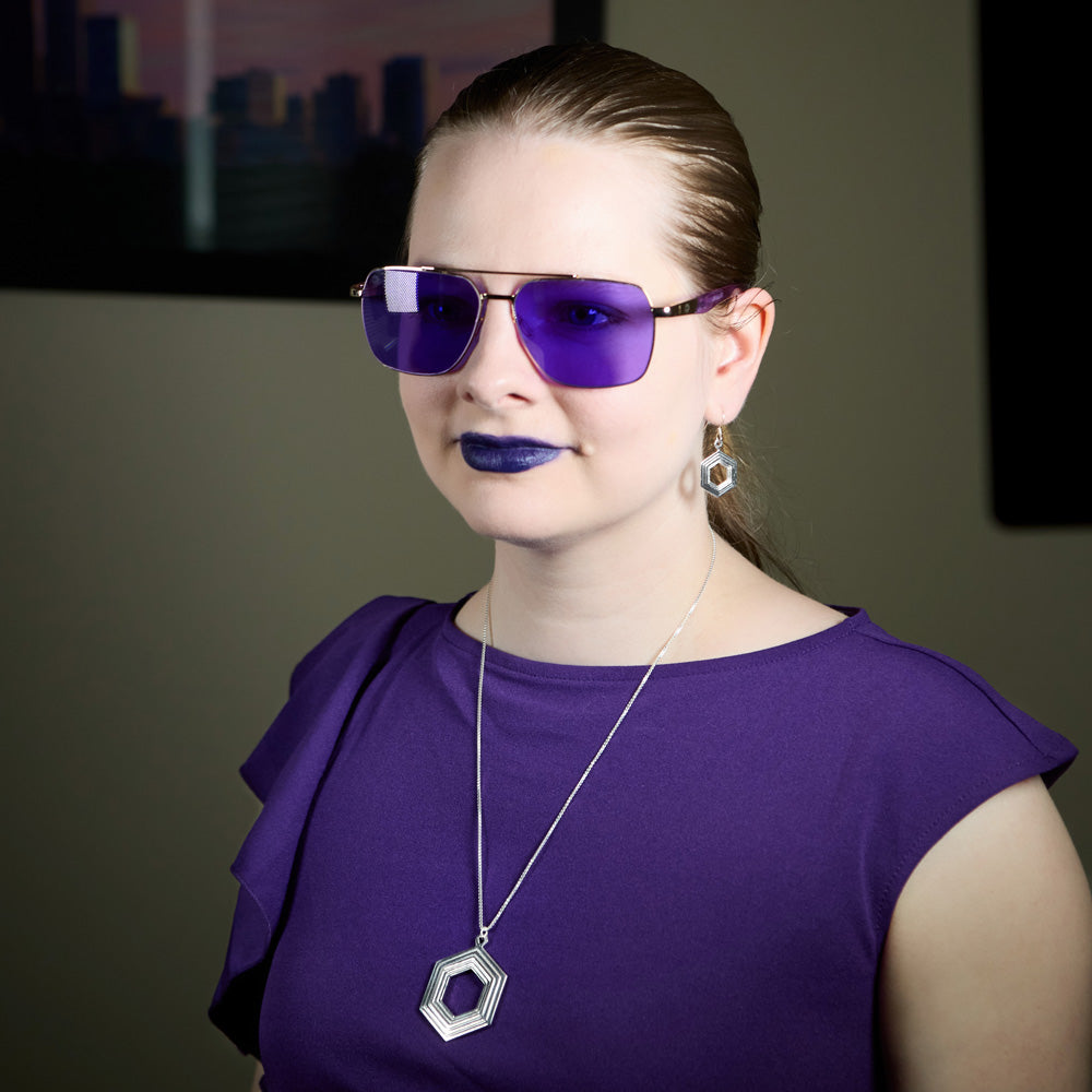 A blonde model wearing our Stepped Hexagon Necklace and Stepped Hexagon Earrings. The model is in a purple dress, wearing purple sunglasses, and has on purple lipstick. The background is blurred. 