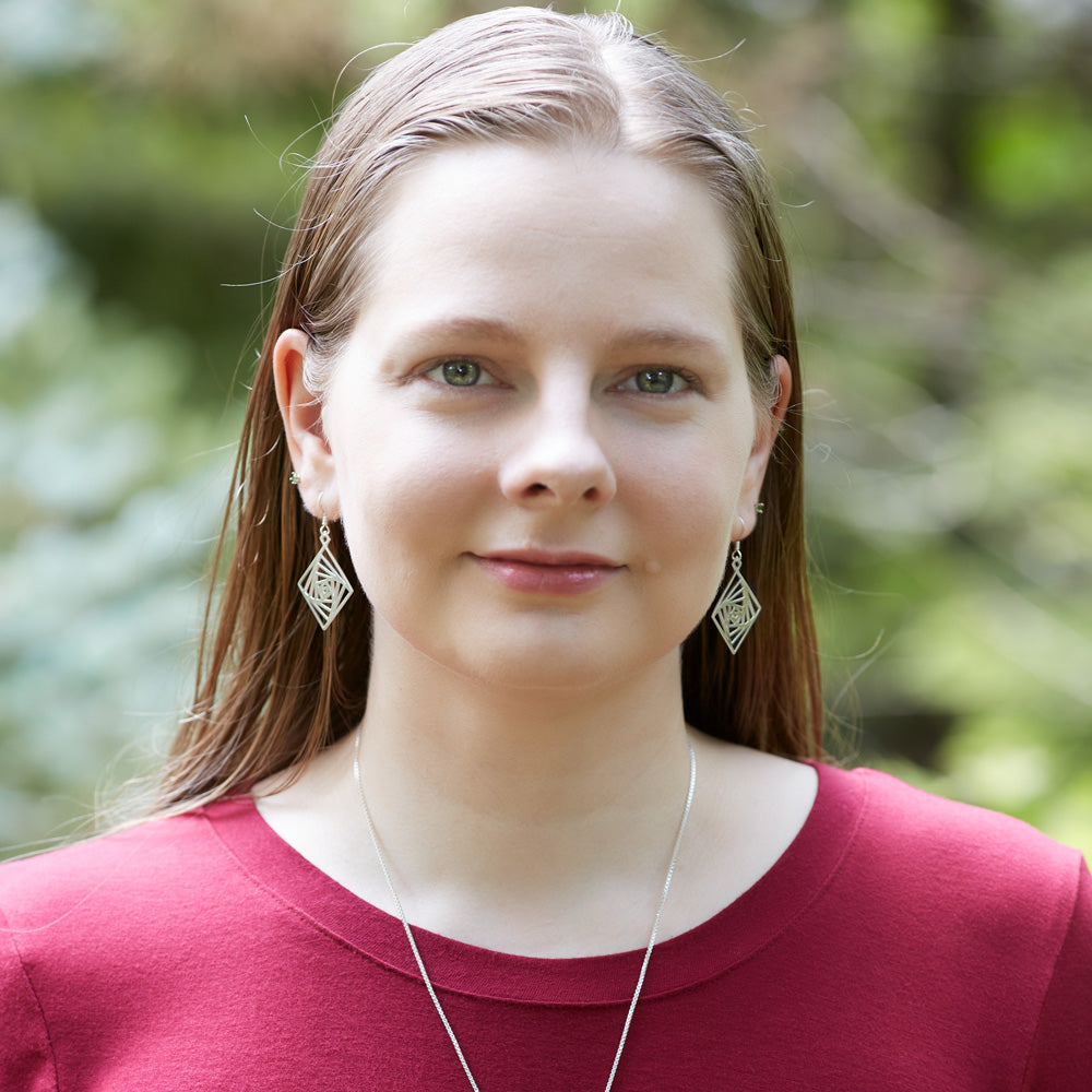 An image of a blonde model wearing the Square Spiral Earrings. The model is looking at the camera and is outdoors. The background is blurred and green. 