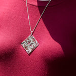 A closeup photo of the Square Spiral Medallion hanging on the neck of a model in a red shirt. The model's face is not visible. The photo is outdoors in the sunlight. 