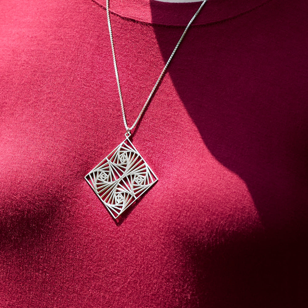 A closeup photo of the Square Spiral Medallion hanging on the neck of a model in a red shirt. The model's face is not visible. The photo is outdoors in the sunlight. 
