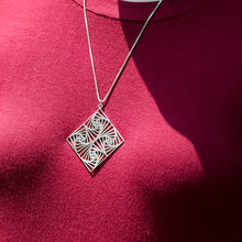Load image into Gallery viewer, A closeup photo of the Square Spiral Medallion hanging on the neck of a model in a red shirt. The model&#39;s face is not visible. The photo is outdoors in the sunlight. 

