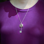 An image of a model wearing the D20 Key Necklace over a purple shirt. The focus is on the pendant, and the photo is taken outdoors. The silver shines in the sunlight. 