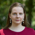 A blonde model wears a pair of Hexwave Earrings outdoors. The model is wearing a red shirt, and is looking at the camera. 