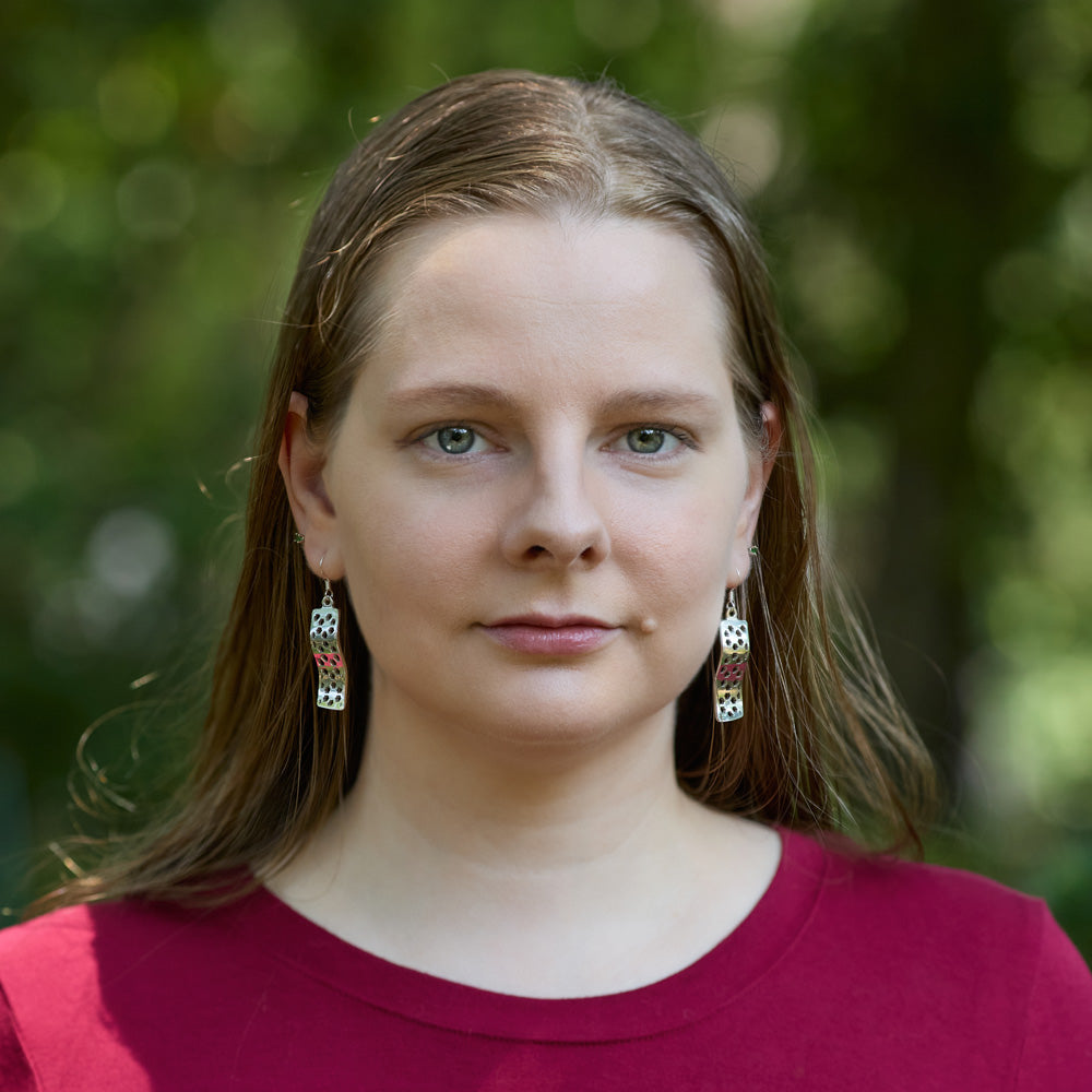 A blonde model wears a pair of Hexwave Earrings outdoors. The model is wearing a red shirt, and is looking at the camera. 