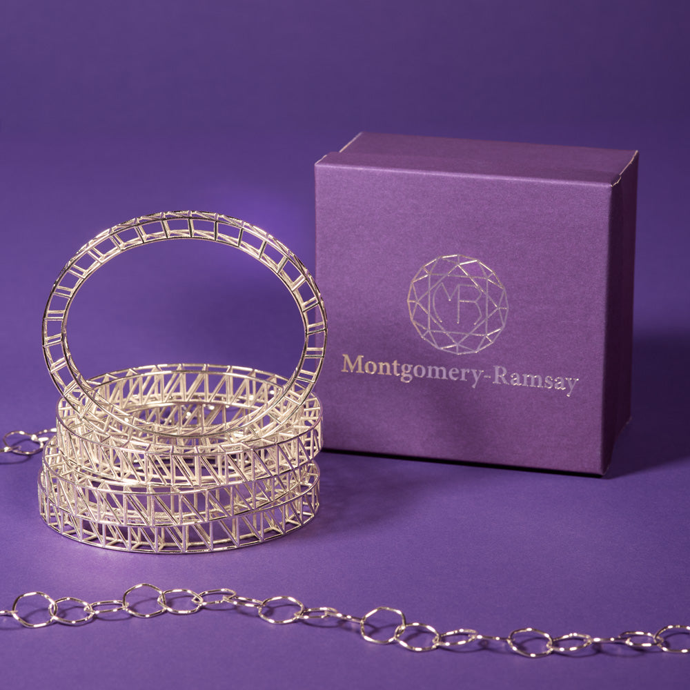A photo of a stack of Scaffold Bangles sitting next to a Montgomery-Ramsay product box. The table and background are both purple, and there is a hexagonal sterling silver chain trailing around in the foreground and background. 