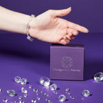 An image of a model's wrist and hand, palm facing the camera above a purple Montgomery-Ramsay product box. The model is wearing a Starlight Prism Bracelet. The background is purple, and there are a collection of clear gems scattered across the purple table. 