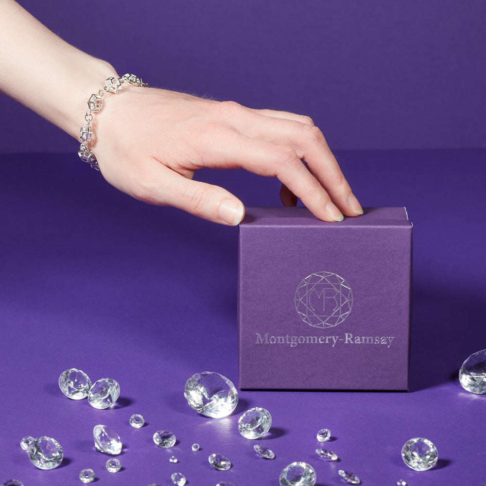 An image of a model's wrist and hand, resting on a purple Montgomery-Ramsay product box. The model is wearing a Starlight Prism Bracelet. The background is purple, and there are a collection of clear gems scattered across the purple table. 