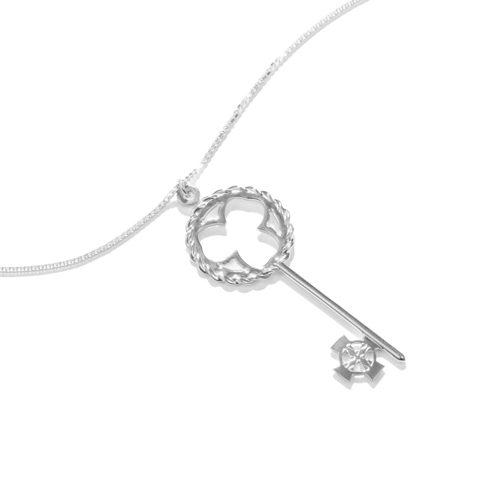 An image of the Celtic Key Necklace at a 45 degree angle, lying on a white surface. The sterling silver Celtic Key Necklace has a bow comprised of a twisted sterling pattern surrounding a classic shamrock design, and a Celtic cross bit and wards. There's a Celtic knot design engraved into the bit and wards to finish off the motif. In very Montgomery-Ramsay fashion, the bail of the necklace has an inset hexagonal design for that small added hallmark detail. 