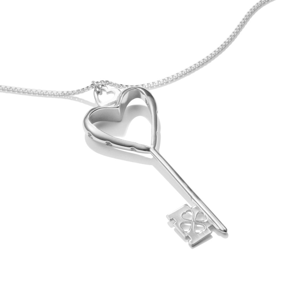 A Heart of Hearts Key Necklace lies in a white surface at a slight angle. The sterling silver key features an elegant heart shape with thickened edges, heart-shaped windows, and is topped with a heart-shaped bail. The wards are also hearts. 