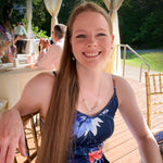A photo of a smiling, young, blonde model wearing our Hexagonal Ice Crystal Necklace. The model is in a flower print dress, sitting at a table amongst other people at an outdoor wedding, and is staring at the camera with a large smile. 