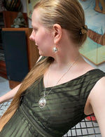 A photo of a model with long blonde hair, leaning back in a chair and wearing our Cross Ball Earrings and our Scrying Pool Necklace. In the background is sculpture and paintings. 