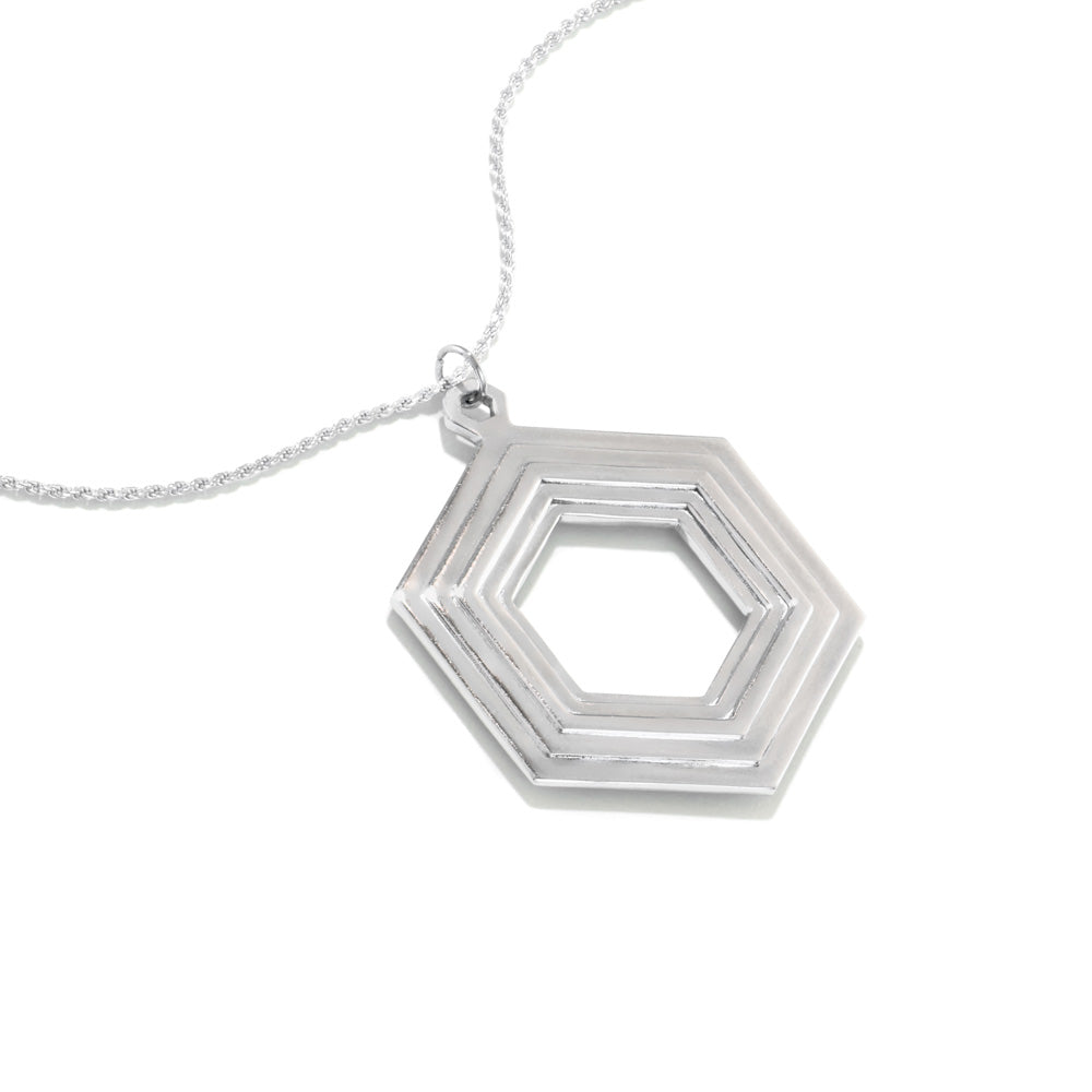 A Stepped Hexagon Necklace lies on a white surface at a 45 degree angle. This large sterling silver necklace is a statement piece, comprised of a modern geometric design featuring rising and falling levels of hexagons. Part of our Stepped Hexagon collection.