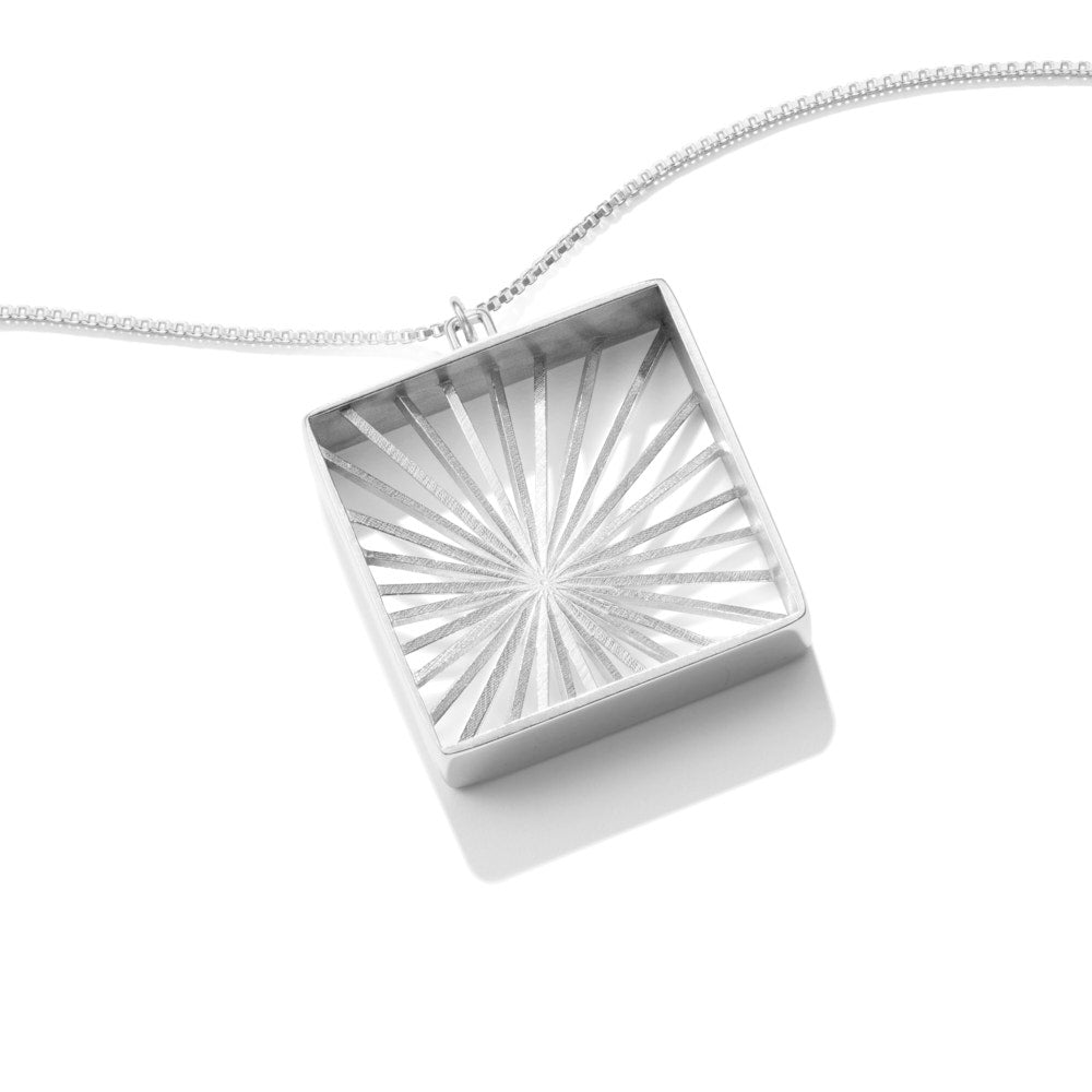 An image of our Sunburst Necklace lying at an angle on a white surface. The Sunburst Necklace is a piece inspired by Barbara Hepworth's stringed figure pieces. Rays of silver sunlight radiate outward from an off-center spot, like bright sunbeams through a window. The piece is 10mm in depth, and a 35mm square (with another 5mm bail). A true statement piece. 