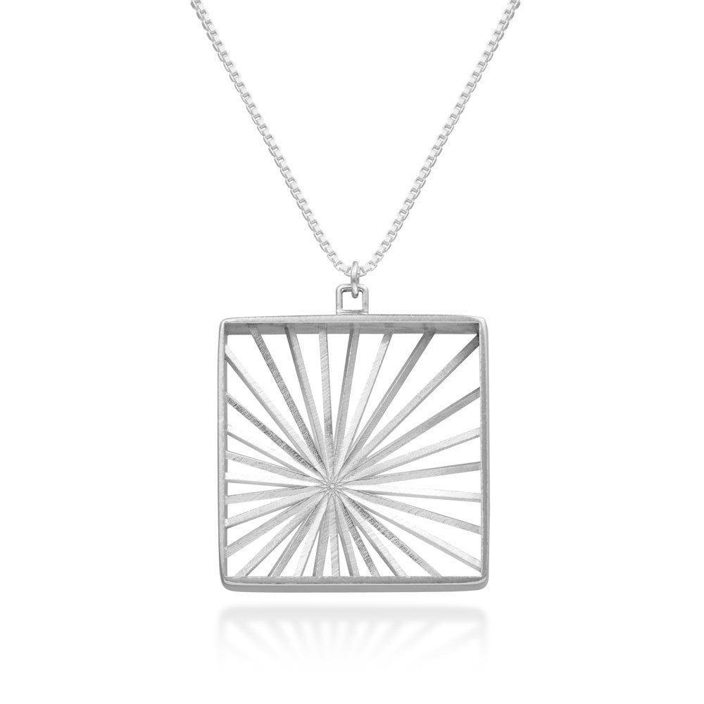 An image of our Sunburst Necklace hanging in front of a white background. The Sunburst Necklace is a piece inspired by Barbara Hepworth's stringed figure pieces. Rays of silver sunlight radiate outward from an off-center spot, like bright sunbeams through a window. The piece is 10mm in depth, and a 35mm square (with another 5mm bail). A true statement piece. 