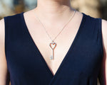 A model in a dark, low-cut navy dress wears a Heart of Hearts Key Necklace. The necklace sparkles in the sunlight. 