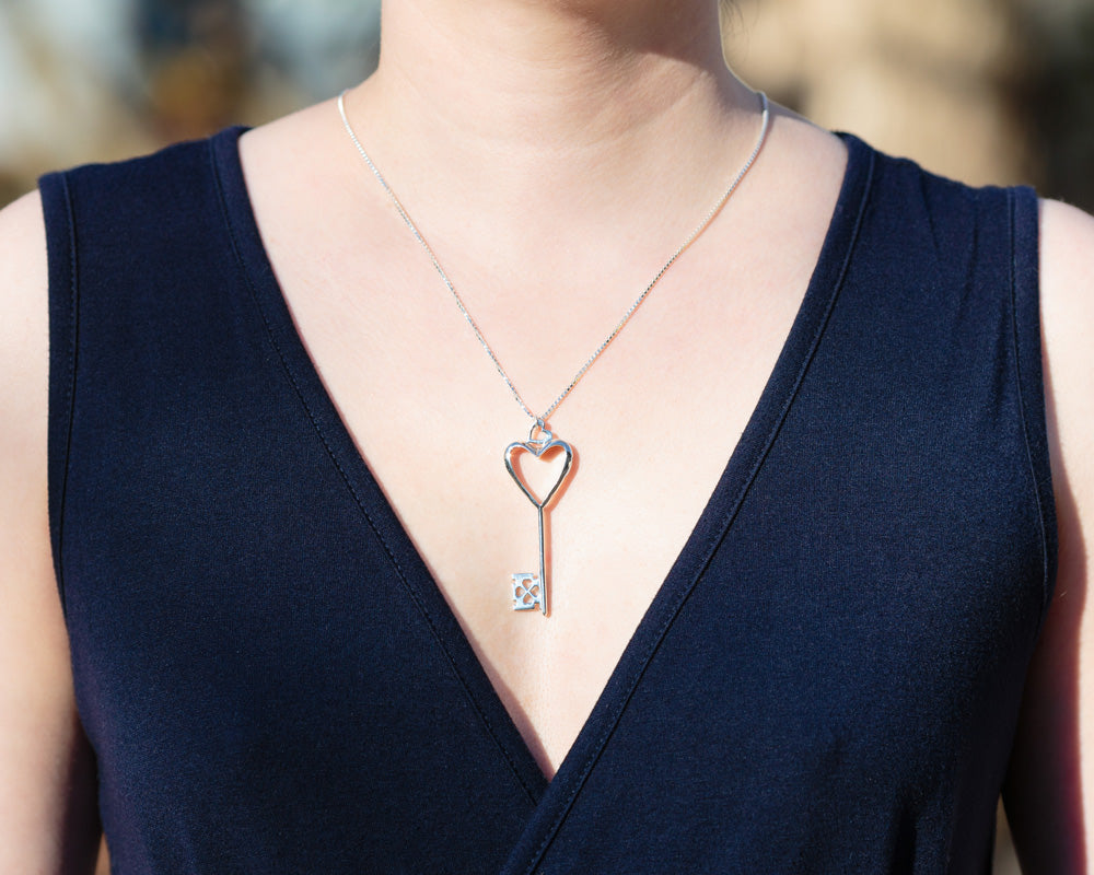 A model in a dark, low-cut navy dress wears a Heart of Hearts Key Necklace. The necklace sparkles in the sunlight. 