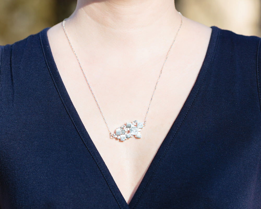 A photo of a model in a navy blue dress wearing our Sea of Squares necklace. The photo is a closeup of the necklace on the model's chest. 