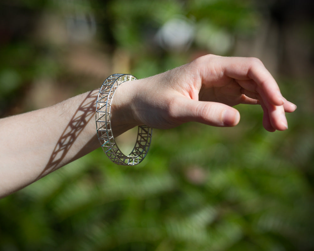 A photo of the wrist of a model with the Scaffold Bangle hanging on it. The model's arm is in sunlight, and the bangle reflects the sun. The background is out of focus. 