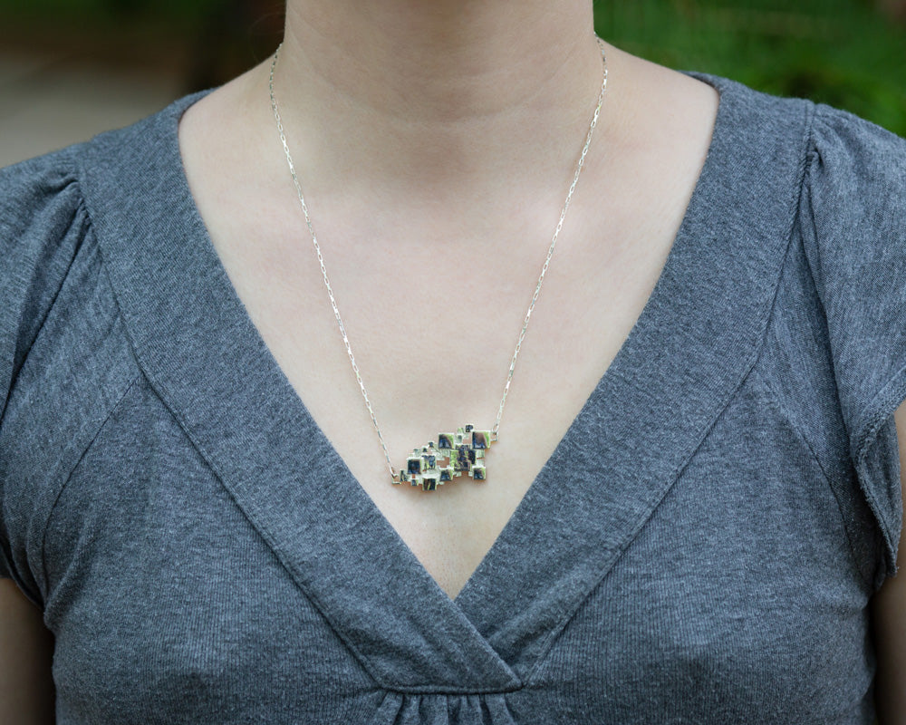 A photo of a model in grey wearing our Sea of Squares necklace. The photo is a closeup of the necklace on the model's chest. 