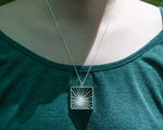 A model wears our Sunburst Necklace outdoors in the sun. The model's face is not shown, and the photo focuses on the necklace in center lower frame. It glints in the sunlight. 