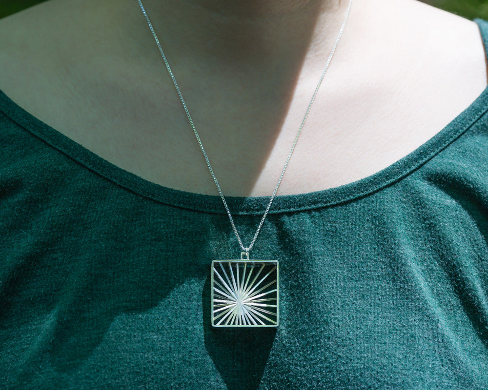 A model wears our Sunburst Necklace outdoors in the sun. The model's face is not shown, and the photo focuses on the necklace in center lower frame. It glints in the sunlight. 