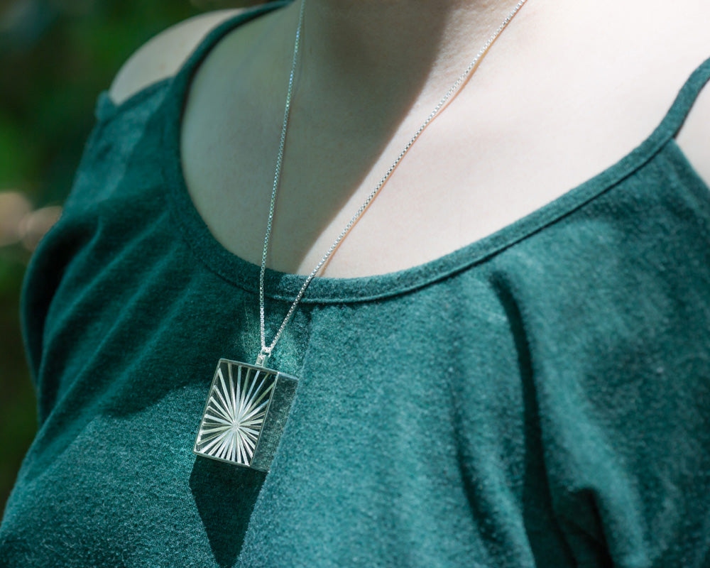 A model wears our Sunburst Necklace outdoors in the sun. The model's face is not shown, and the photo focuses on the necklace in center lower frame. It glints in the sunlight, and this view shows a view from the front left of the necklace. 