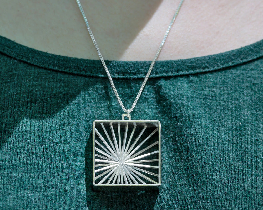 A model wears our Sunburst Necklace outdoors in the sun. The model's face is not shown, and the photo focuses closely on the necklace in center lower frame. It glints in the sunlight. 