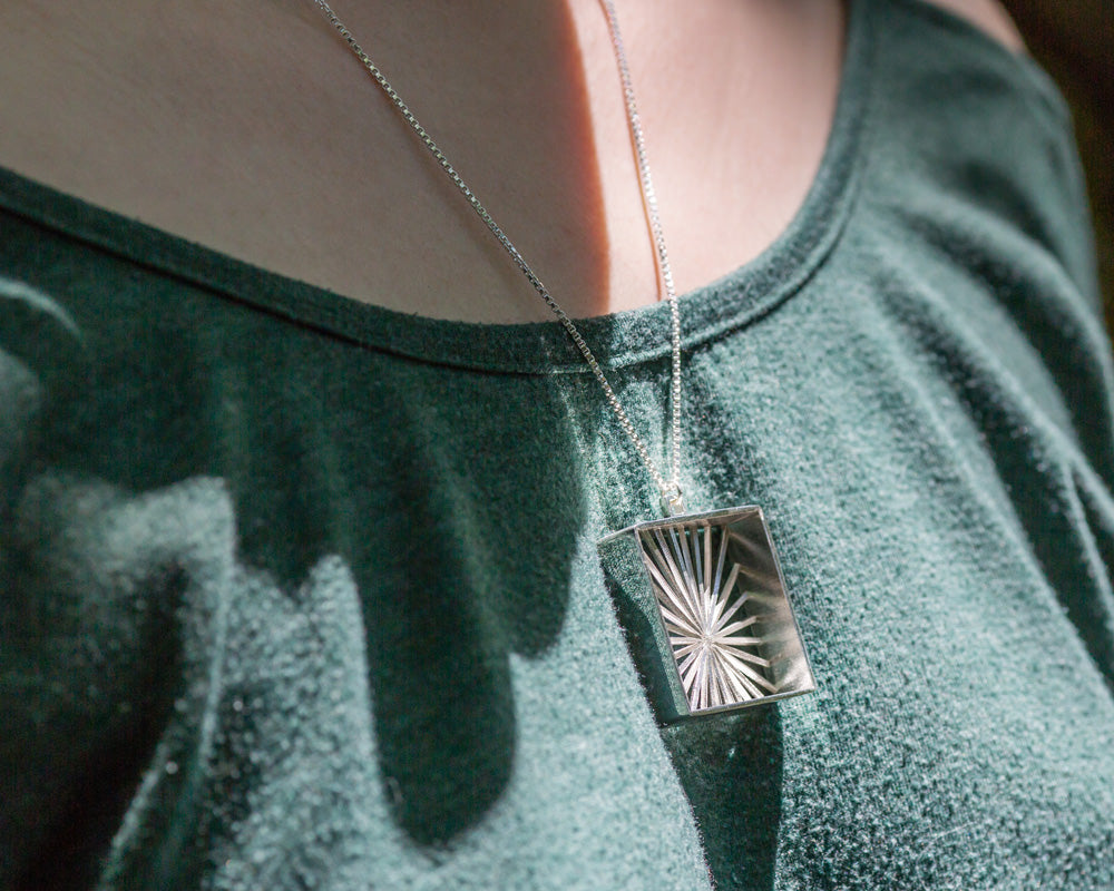 A model wears our Sunburst Necklace outdoors in the sun. The model's face is not shown, and the photo focuses on the necklace in center lower frame. It glints in the sunlight, and this view shows a view from the front right of the necklace. 