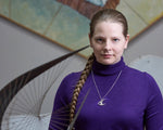 A blonde model in a purple turtleneck wears a Curlew Necklace Pendant. The model is looking at the camera. In the foreground, slightly blurred, there is the Barbara Hepworth Curlew sculpture that inspired this design. There is art in the background, but blurred. 