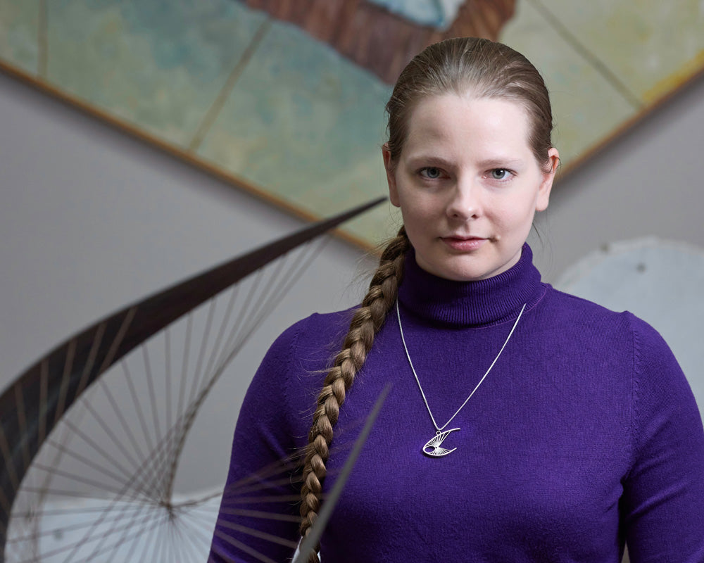 A blonde model in a purple turtleneck wears a Curlew Necklace Pendant. The model is looking at the camera. In the foreground, slightly blurred, there is the Barbara Hepworth Curlew sculpture that inspired this design. There is art in the background, but blurred. 